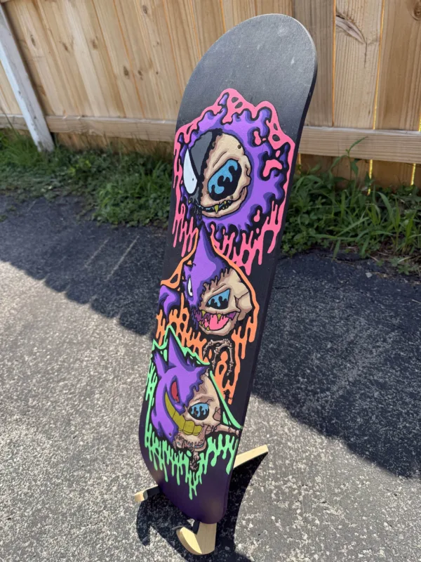 Side view of the hand-painted skateboard deck with Gastly, Haunter, and Gengar skulls. Acrylic gloss finish catches the light, emphasizing vivid neon tones and dripping textures.