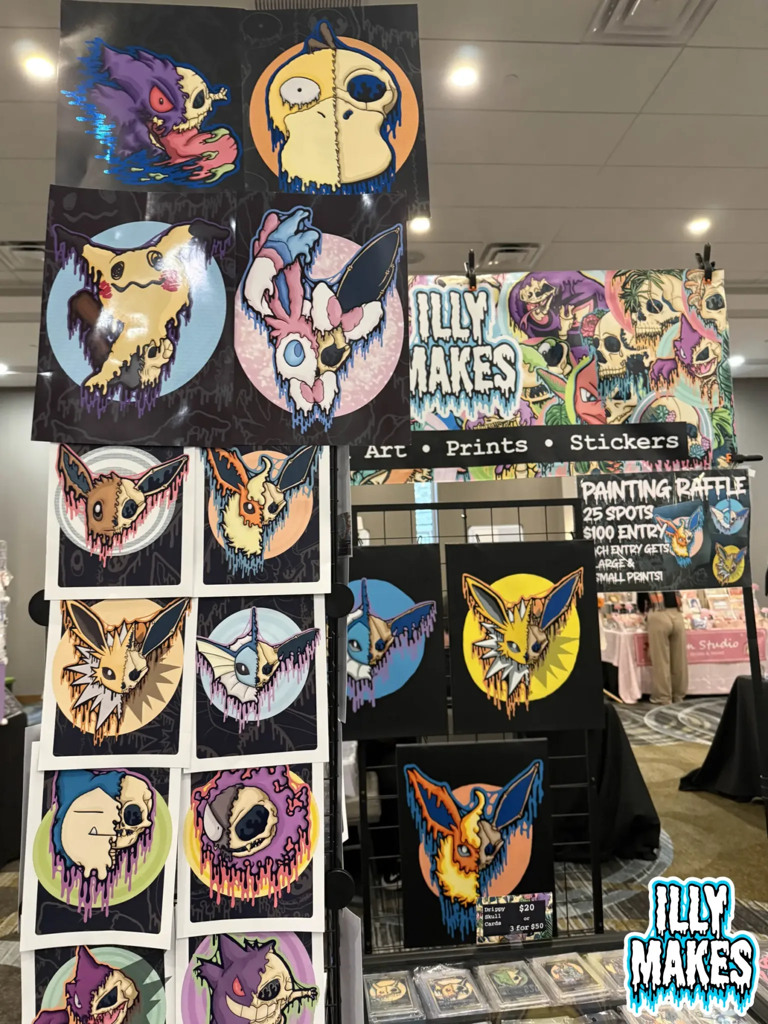 Close-up view of my merch table featuring drippy skull prints, Pokémon skull stickers, and original canvas paintings.